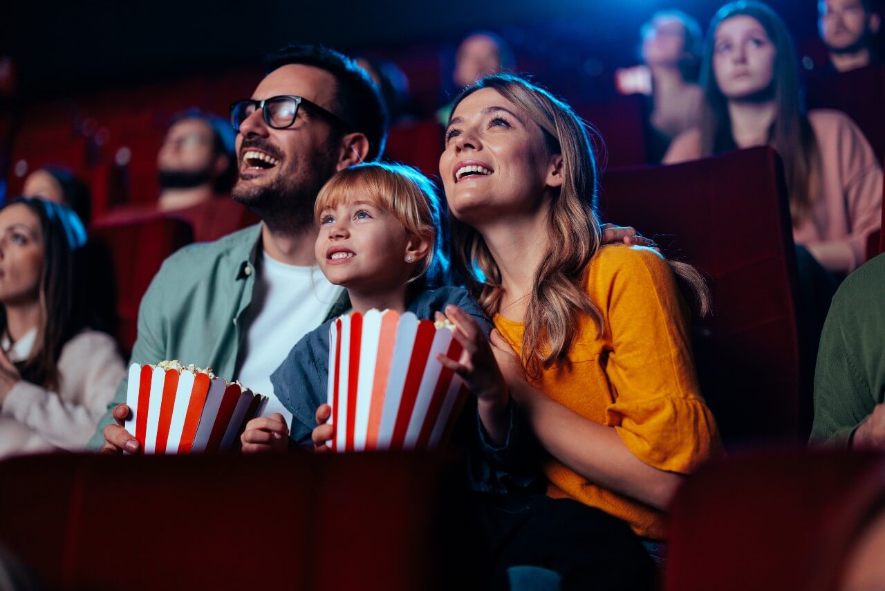 parents and a kid looking happily at the screen at a crowed movie theatre holding 2 buckets of popcorn.