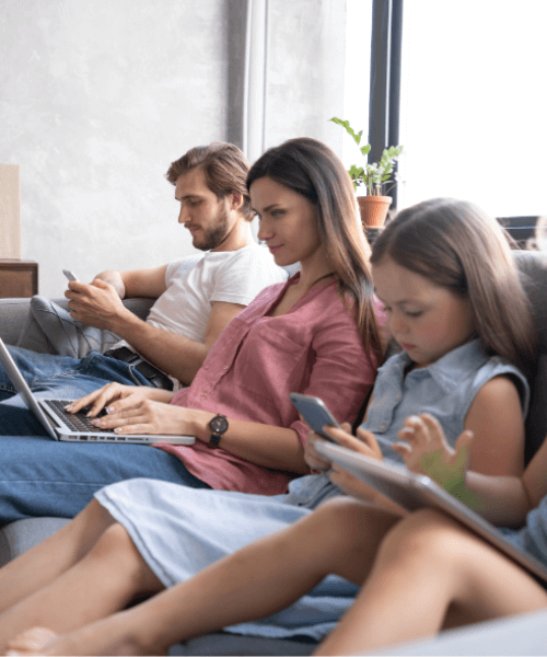 Family sitting on a couch. Parents on their phone and laptop and 2 kids on their phone and tablet